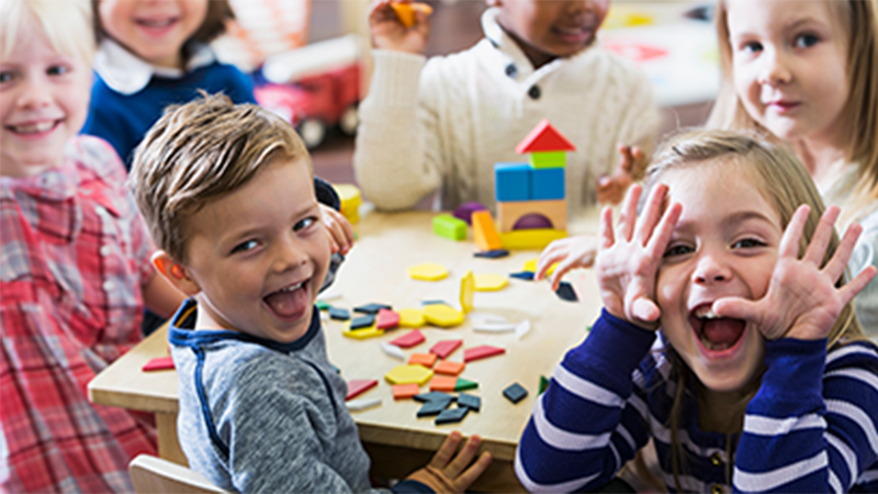 A group of very young children smiling and laughing together at a table with colorful, wooden building blocks