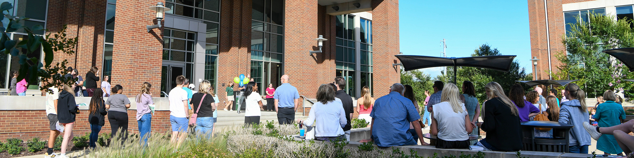 A crowd of adults gathered outside a brick building with large windows, some sitting on benches, as others stand in line.