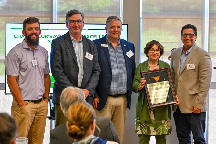 A group of five people stand in front of a window while one person holds a framed award.