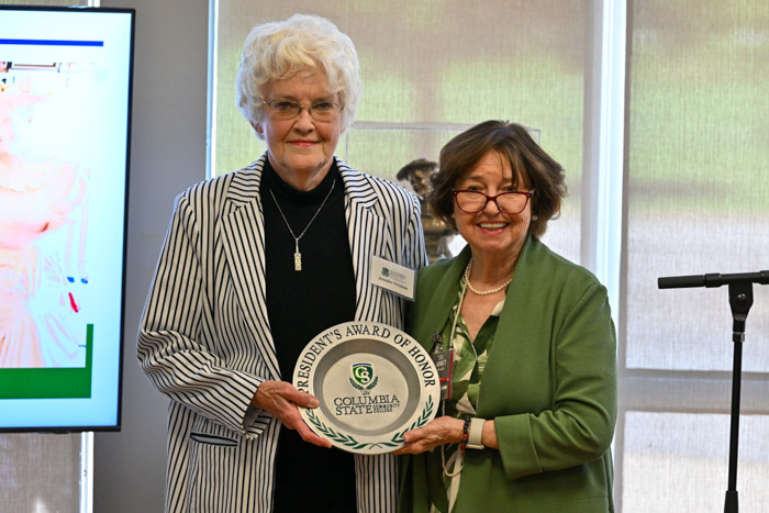 Two people smile for a photo. The person on the left is holding a plate-shaped award.