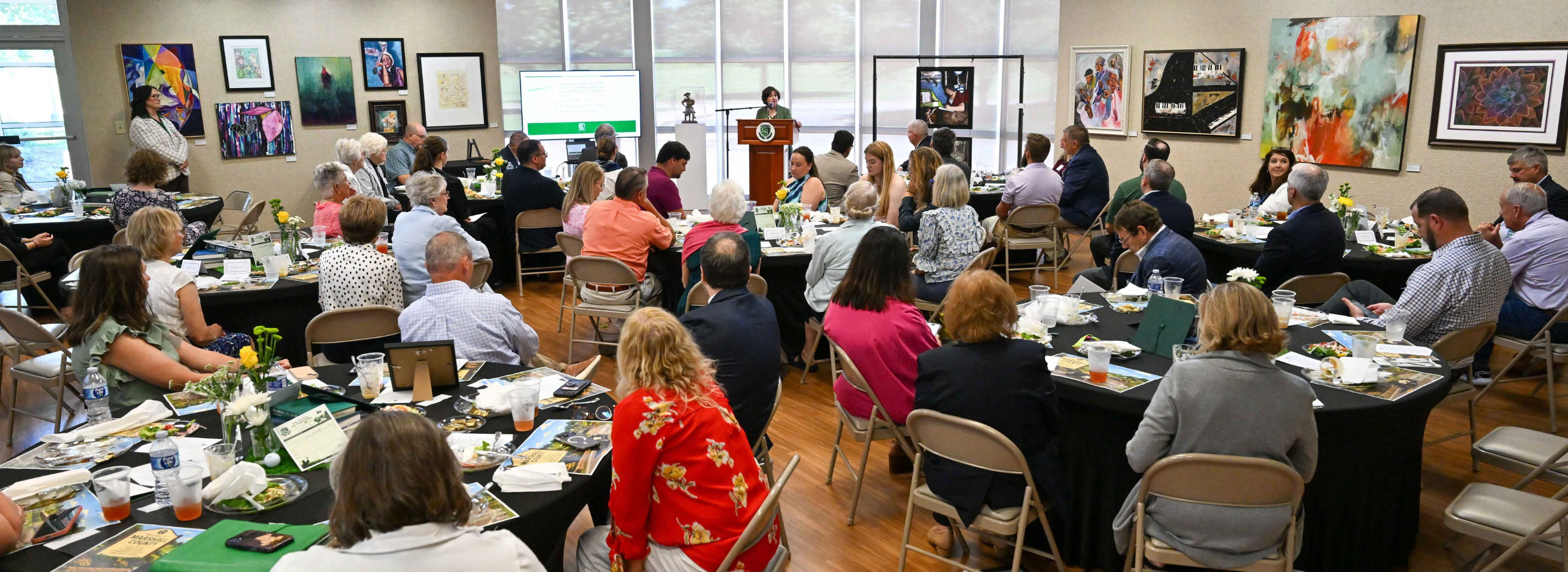 Groups of people are seated at tables eating while a person speaks at a podium.