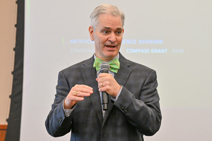A person speaks into a microphone at the front of a room while wearing a gray suit and green bow tie.