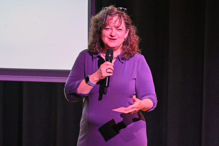 A person wearing a purple dress speaks on a stage while using a microphone.
