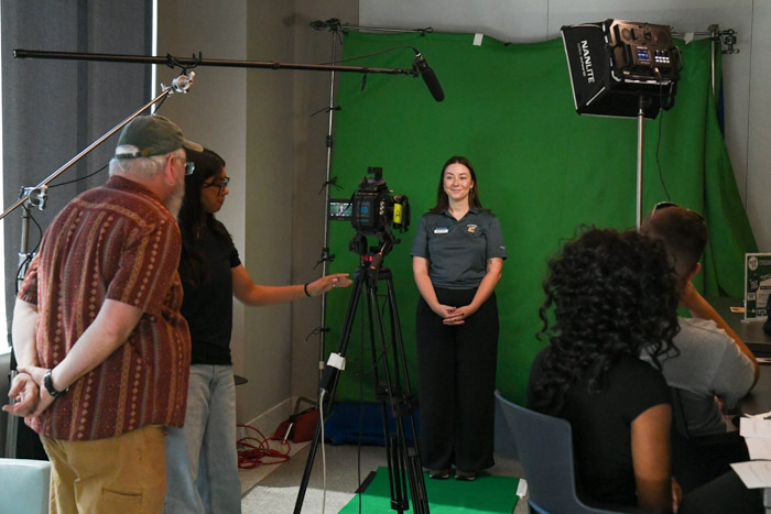 A person stands in front of a green screen while people stand behind a video camera.